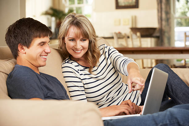 Smiling mother using laptop with teenage son on the couch Mother With Teenage Son Sitting On Sofa At Home Using Laptop mother and teenage son stock pictures, royalty-free photos & images