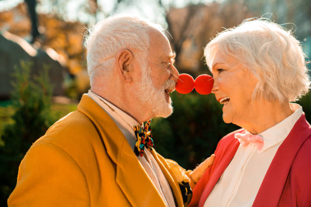 Smiling old husband and wife with clown noses in the park Side view of happy elderly couple in colorful costumes and clown accessories clown's nose stock pictures, royalty-free photos & images