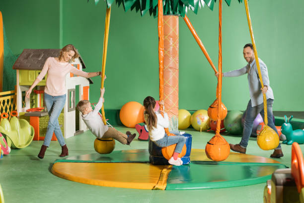 smiling parents looking at happy children swinging on swings in entertainment center smiling parents looking at happy children swinging on swings in entertainment center indoor playground stock pictures, royalty-free photos & images