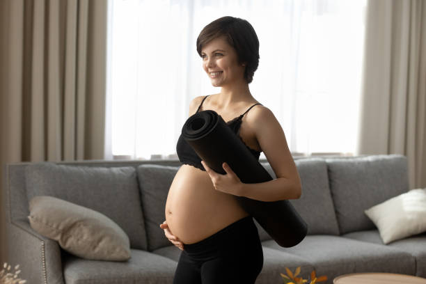 Smiling pregnant woman in black sportswear holding yoga mat Smiling pregnant woman in black sportswear holding yoga mat, touching belly, happy young future mom ready for training, gymnastics or exercise during pregnancy, healthy lifestyle and sport pregnant weight stock pictures, royalty-free photos & images