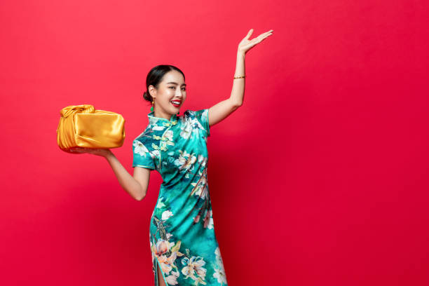 Smiling pretty Asian woman in oriental style costume with golden gift box in red isolated studio background for Chinese new year concepts stock photo