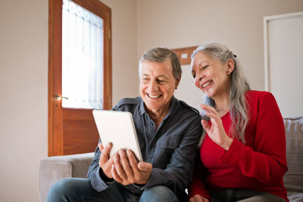 Smiling senior couple using dibital tablet Cheerful senior woman and man sitting on sofa at home and having video conference, using digital tablet. Lovely elderly female and male are laughing in living room. social work online stock pictures, royalty-free photos & images