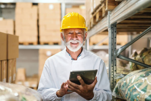 glimlachend senior werknemer in uniform en met gele helm op het hoofd houden van de tablet pc- en kijken naar camera terwijl je in opslag. - pensioen nederland stockfoto's en -beelden