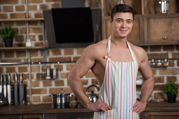 smiling sexy shirtless man standing in apron at kitchen - wearing nothing b...