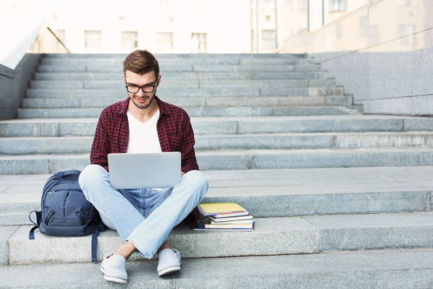 Smiling student sitting on stairs using laptop Smiling student sitting on stairs working with laptop, preparing for exams outdoors, having rest in university campus. Technology, education and remote working concept, copy space universities in europe for computer science stock pictures, royalty-free photos & images