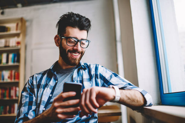 Smiling successful hipster guy in eyewear checking time on wearable smartwatch while waiting for meeting at university campus, happy man holding mobile phone in hand and reading message on wristwatch stock photo