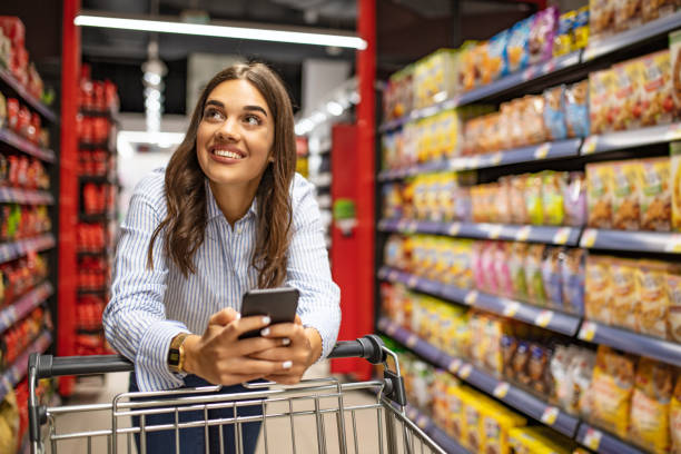 Smiling woman at supermarket. Smiling woman at supermarket. Happy woman at supermarket. Beautiful young woman shopping in a grocery store/supermarket. Shopping lists in app format grocery store stock pictures, royalty-free photos & images