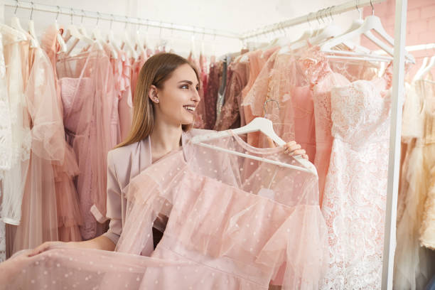Smiling Woman Choosing Dress in Boutique Waist up portrait of smiling beautiful woman choosing feminine dress while standing against clothing racks in shop, copy space evening gown stock pictures, royalty-free photos & images