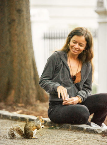 Smiling woman sitting on a footpath curb in a public park and feeding nuts to a cute little squirrel