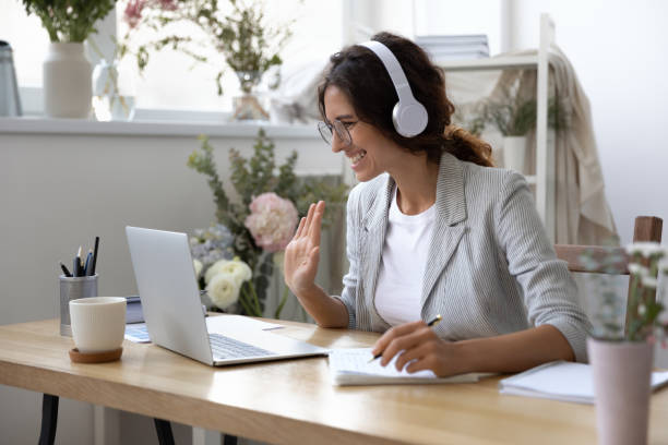Smiling woman in headset have inline educational course Smiling young Caucasian woman in headphones take online educational course or training on laptop from home, happy female in wireless headset wave to camera, talk on webcam video call on computer virtual reality stock pictures, royalty-free photos & images