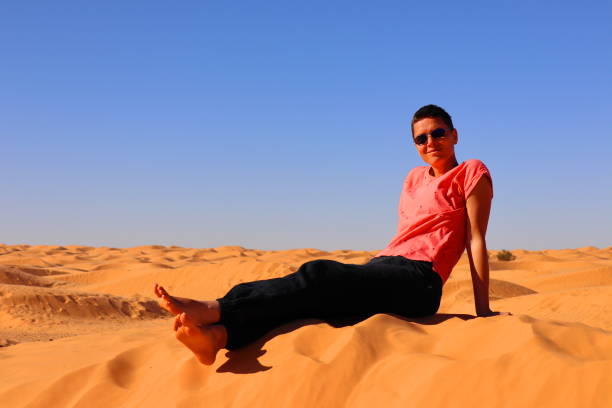 Smiling woman sitting on the sand dunes with blue sky Female traveler with short hair and sunglasses enjoying the sahara desert near Douz, Tunisia on a sunny day with clear sky. tunisia woman stock pictures, royalty-free photos & images