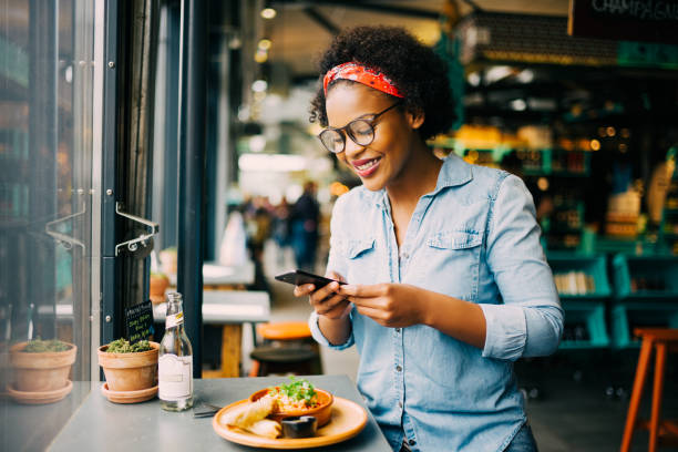 Smiling woman taking photos of her food in a cafe Young African woman smiling while sitting alone at a counter in a bistro taking photos of her meal with her smartphone eating photos stock pictures, royalty-free photos & images
