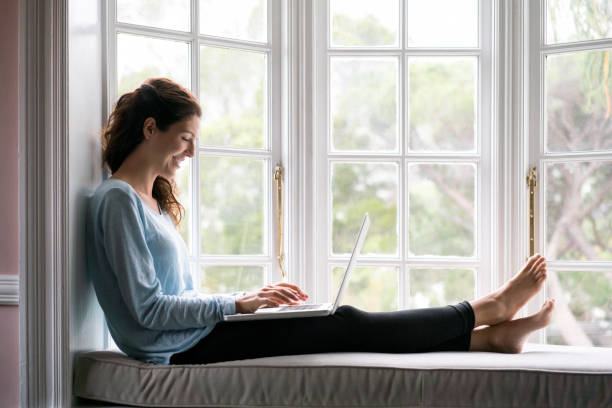 Smiling woman using laptop at alcove window seat Smiling woman using laptop while sitting at alcove window seat. Full length of female is wearing casuals. She is at home. alcove window seat stock pictures, royalty-free photos & images