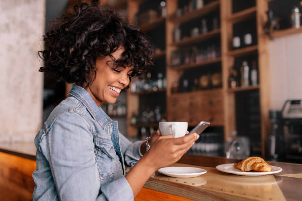 Smiling woman using smart phone in a modern cafe Smiling woman using smart phone in a modern cafe. Young african female sitting at a cafe counter having coffee and reading text message on mobile phone. curley cup stock pictures, royalty-free photos & images