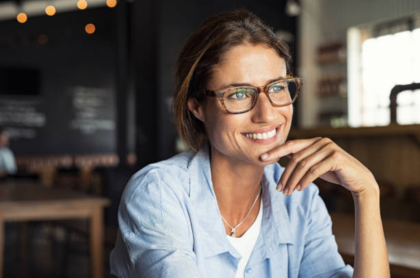 Smiling woman wearing spectacles Portrait of beautiful mature woman sitting in cafeteria looking away. Cheerful mature woman wearing eyeglasses thinking with finger on chin. Happy woman relaxing at cafe and smiling. day dreaming photos stock pictures, royalty-free photos & images