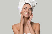 Studio portrait over grey perfect woman with white towel on head after shower having toothy smile touch gently healthy shiny clean skin looking at camera, skincare and natural beauty treatment concept