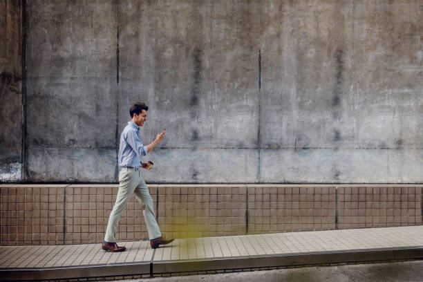 Smiling Young Asian Businessman in Casual wear Using Mobile Phone while Walking by the Urban Building Wall stock photo