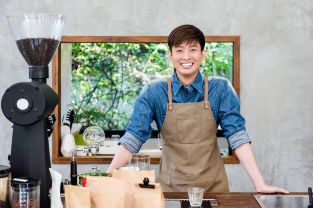 Smiling young Asian man entrepreneur at coffee shop counter bar Cheerful smiling young Asian man entrepreneur standing at coffee shop counter ready to welcome and service customer bar drink establishment photos stock pictures, royalty-free photos & images