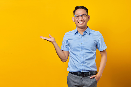 smiling young asian man in glasses; showing copy space on palm isolated on yellow background.