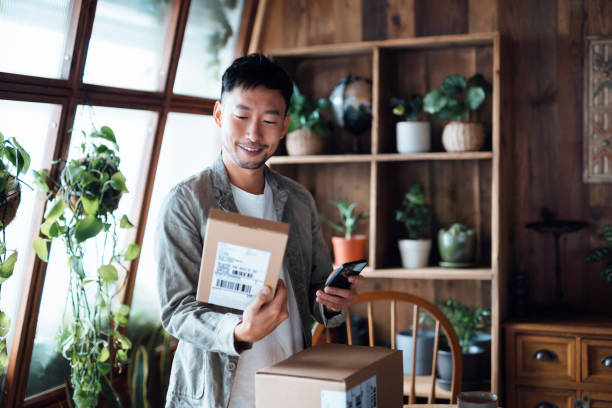 smiling young asian man with smartphone, receiving delivered packages from online purchases at home, can't wait to unbox the purchases. online shopping, enjoyable customer shopping experience - unbox stockfoto's en -beelden