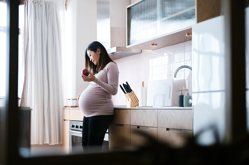 Smiling young Asian pregnant woman touching her belly, eating an fresh organic apple in kitchen at home. Healthy diet. Eating well. Healthy pregnancy eating lifestyle