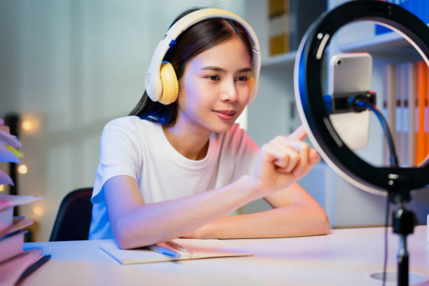 Smiling Young Asian woman wearing headset and live broadcasting on internet and reading comments with people in social media on smartphone. stock photo