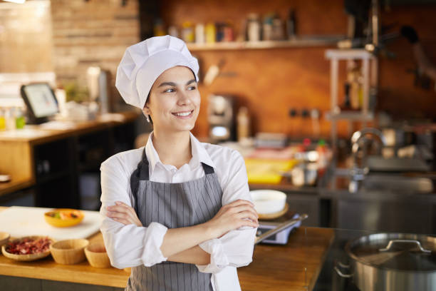 Smiling Young Cook wearing Chefs Hat Waist up portrait of cheerful young cook posing confidently and looking away while standing in restaurant kitchen, copy space chef stock pictures, royalty-free photos & images
