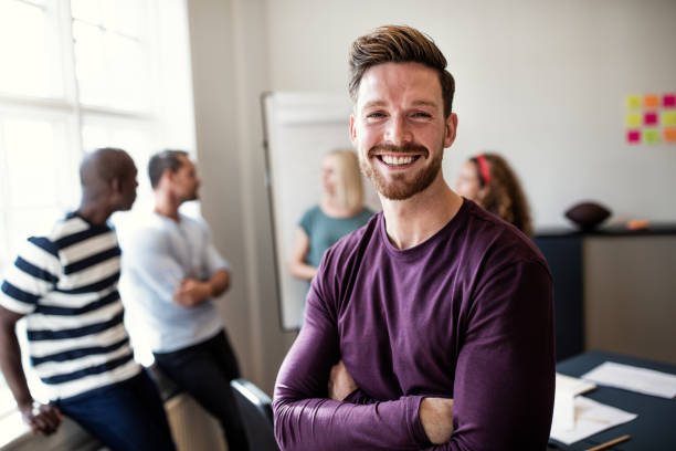 Smiling young designer standing in an office after a presentation Smiling young designer standing confidently with his arms crossed after a boardroom meeting with colleagues standing in the background scandinavia photos stock pictures, royalty-free photos & images