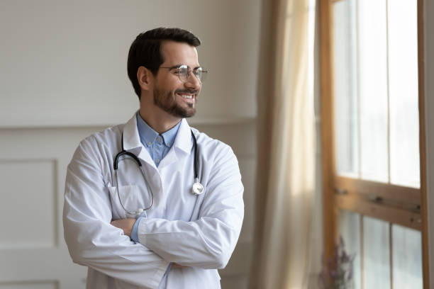 Smiling young doctor planning future career, standing near window. Smiling young doctor in white medical uniform standing with folded hands near window, lost in thoughts. Positive handsome male head of department thinking of challenges, planning future career. dermatologist stock pictures, royalty-free photos & images