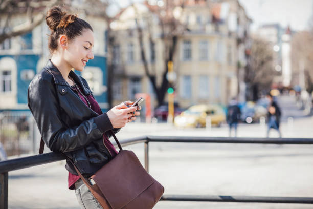 Smiling young female browsing on her phone while waiting on the street stock photo