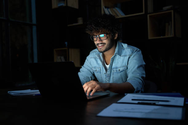Smiling young indian business man working online late at night using laptop. stock photo