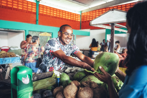 smiling young market vendor giving fresh coconut to female client at market stall at public market in Bahia, Brazil