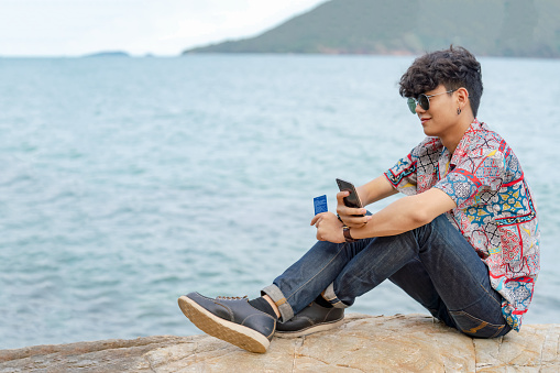 Smiling young smart Asian man guy sitting on rocky seacoast beach using smartphone with internet for online shopping and credit card for e-banking payment. E-commerce and wireless technology concept