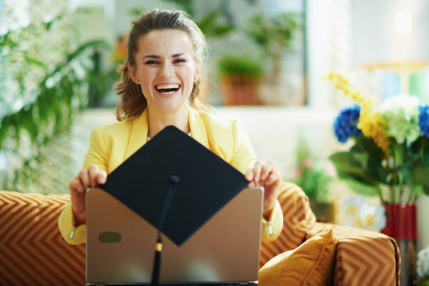 smiling young student showing graduation cap and laptop smiling young student in jeans and yellow jacket at modern home in sunny day showing graduation cap and laptop. degree online stock pictures, royalty-free photos & images
