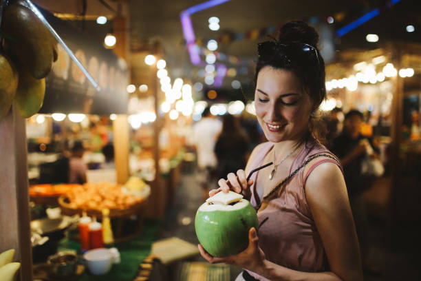 Smiling young woman having a delicious and fresh coconut water on the go Authentic Thai street food in Bangkok. Young woman is having a refreshment with delicious coconut water on a hot summertime evening. night market stock pictures, royalty-free photos & images