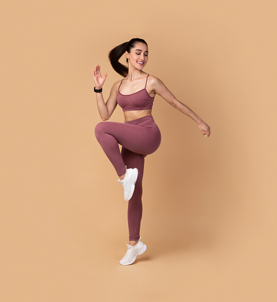 Training Concept. Full Length Portrait Of Smiling Fit Young Lady In Sportswear Jumping And Lifting Leg Up, Exercising Isolated On Pastel Background. Studio Shot, Free Space. Energy And Sports
