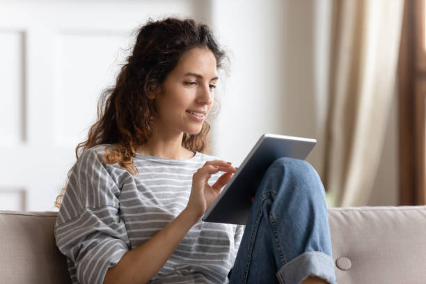 컴퓨터 태블릿을 사용하여 미소 짓는 젊은 여자, 소파에 앉아 - 디지털 태블릿 사용하기 뉴스 사진 이미지