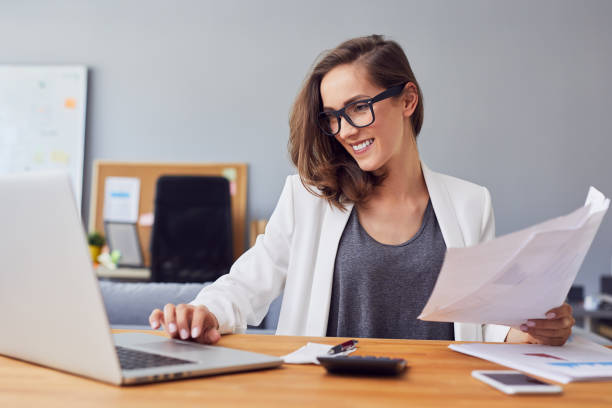 Smiling young woman working in home office using laptop and documents Smiling young woman working in home office using laptop and documents accounting ledger stock pictures, royalty-free photos & images