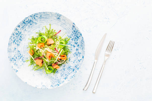 Smoked salmon salad with Arugula, apple, enoki mushrooms, zucchini and beetroot on a white and blue plate aside cutlery over a blue background with copy space. stock photo