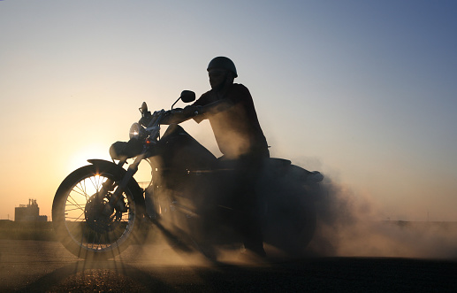 Silhouette of motorcycle and rider against blue prairie sky. Sunlight casts gorgeous shadows through wheel spokes, while smoke from burning tire billows behind bike