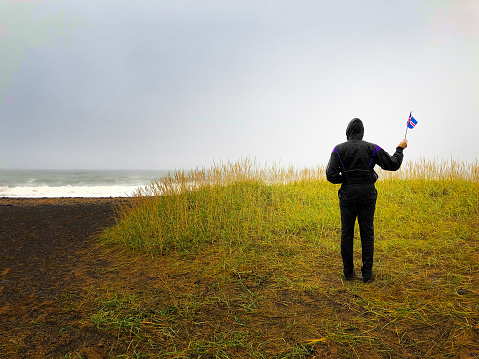 Snaefellsnes, Iceland: A man in black raingear waving an Icelandic flag on a  beach in Western Iceland in autumn.