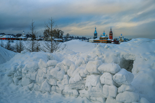 Children from the snow built a snow fortress for their fun. Old churches and wooden houses of the provincial town of Cherdyn (Northern Urals, Russia) are visible in the distance. Winter evening