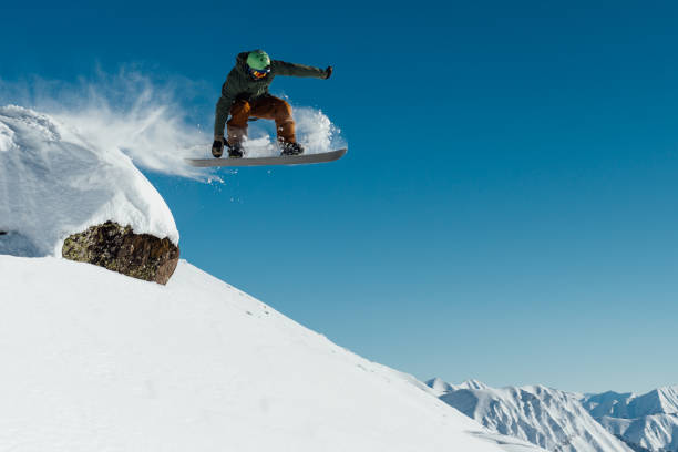 snowboarder in the outfit drops off the ledge of the stone onto the fresh snow creating a spray of snow stock photo