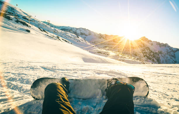 snowboarder sitting at sunset on relax moment in french alps ski resort - winter sport concept with adventure guy on top of mountain ready to ride down - legs view point with teal and orange filter - snowboard imagens e fotografias de stock