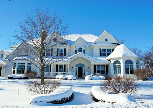A snow covered mansion on a sunny day with blue sky and bare trees.