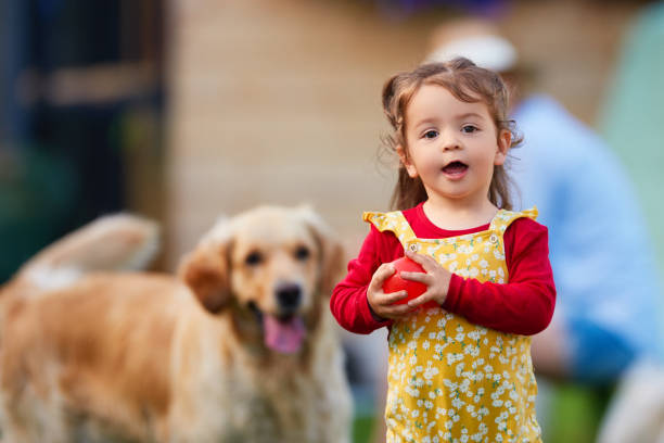 So much fun to play in my garden Portrait of sweet little girl wearing a red T-shirt and a yellow dress outdoors, playing, carrying red ball, dog in the background beautiful young brunette girl playing with her dog stock pictures, royalty-free photos & images