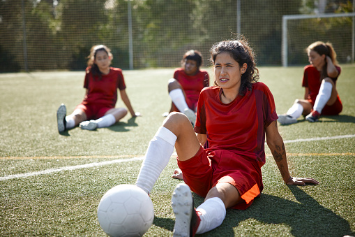 Hispanic female soccer player relaxing with teammates on grass field after practice