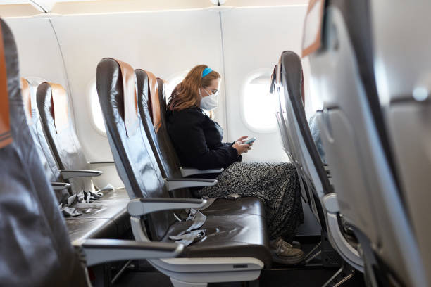 Socially Distanced Portrait of Teenage Traveler in KN95 Mask Side view of solitary Caucasian 17 year old girl wearing casual clothing and protective mask checking smart phone while flying on airplane in time of COVID-19. plane window seat stock pictures, royalty-free photos & images