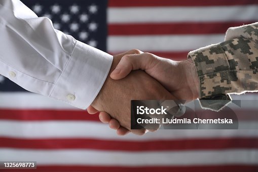 istock Soldier and businessman shaking hands against flag of USA, closeup 1325698773