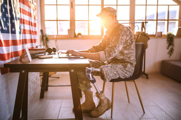 Soldier of American army with prosthetic limb using laptop Young Soldier in uniform with amputee leg sitting on desk in office chair, using lap top military lifestyle stock pictures, royalty-free photos & images